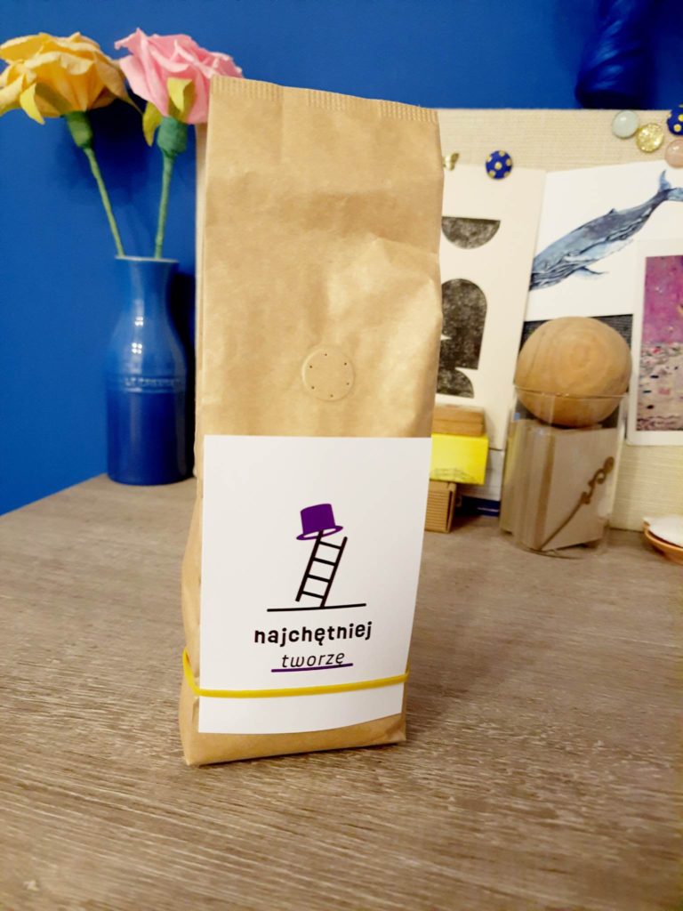Opakowanie kawy - brązowa torebka, do której przyczepiona jest żółtą gumką recepturką etykieta kawiarni wydrukowana na grubszym papierze. 